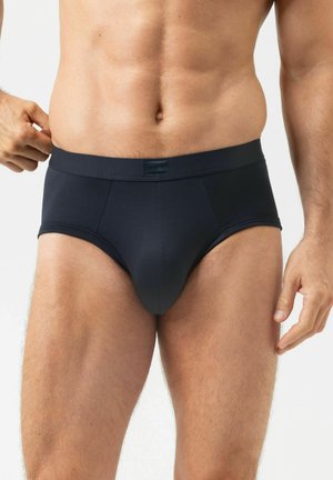 Buste masculin portant un slip noir moulant, montrant un abdomen et des jambes toniques sur un fond clair uni.