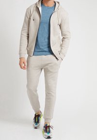 Beige Sweatshirt mit Kapuze, blauem T-Shirt darunter, passenden beigen Jogginghosen und mehrfarbigen Sneakers mit roten Schnürsenkeln sowie grauen und gelben Akzenten.