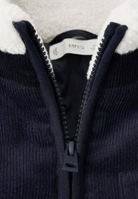 Veste zippée bleu marine avec une texture en velours côtelé et un col moelleux blanc. Comprend une fermeture éclair noire et une étiquette portant la mention ‘MNG’ à l'intérieur.