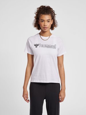 Vrouw met krullend haar die een wit Hummel T-shirt met korte mouwen en een zwarte broek draagt, staand tegen een effen lichte achtergrond.