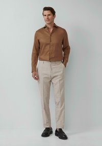 Braunes Leinenhemd mit durchgehender Knopfleiste, kombiniert mit hellbeigen Hosen; schwarze Loafer; einfarbiger Hintergrund.