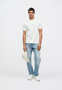 Hvit bomulls-T-skjorte med liten trykt tekst, lyseblå jeans, kremfarget baseballcaps holdt i hånden, og hvite joggesko. Enkel design, avslappet passform.