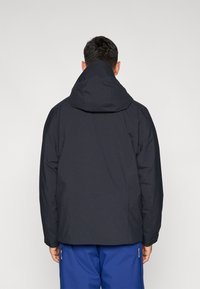 Casaco impermeável preto com capuz, apresentando uma textura suave e design ajustado. Nas costas tem detalhes em costura e punhos ajustáveis.