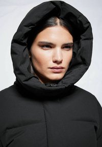 Schwarze gepolsterte Jacke mit einer großen, strukturierten Kapuze, Reißverschluss und glatter Textur, die über genähte Einsätze für gestalterische Details verfügt.