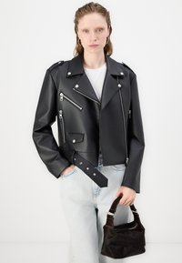 Chaqueta de moto corta de cuero sintético negro que presenta herrajes plateados, bolsillos con cremallera y una cintura con cinturón, combinada con denim claro y un bolso oscuro.