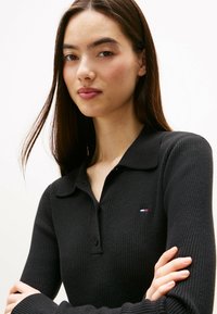 Młoda kobieta z długimi ciemnymi włosami, ubrana w czarną, prążkowaną bluzkę z długim rękawem, z małym logo w czerwono, białym i niebieskim kolorze na piersi.