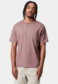 T-shirt di cotone a maniche corte color malva con tasca sul petto sinistro e patch con logo, abbinato a pantaloni chiari.
