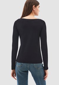 Maglietta nera a maniche lunghe con scollatura ampia, realizzata in tessuto morbido, caratterizzata da una vestibilità aderente e cuciture minimali. Abbinata a jeans blu.