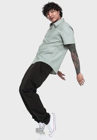 Mintgrünes Hemd mit Knöpfen, dunkle Cargo-Hosen und weiße Sneaker. Das Hemd hat einen Kragen und eine Brusttasche; Tattoos sind am Arm sichtbar.