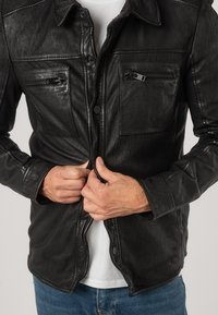 Giubbotto di pelle nera con chiusura a bottoni, due tasche sul petto con cerniera, superficie testurizzata e colletto. Abbinato a una camicia bianca sottostante.