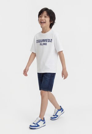 Χαμογελαστό αγόρι που περπατά, φοράει λευκό T-shirt Dsquared2 Milano, σκούρο τζιν σορτς και λευκά-μπλε αθλητικά παπούτσια σε λευκό φόντο.