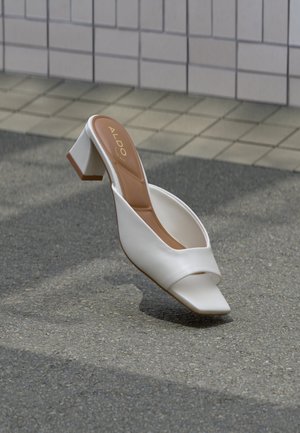 White open-toe mule with block heel and tan insole labeled "ALDO," positioned on asphalt with tiled wall background.
