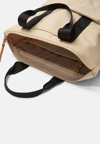 Mochila de lona beige con asas de tela negra, que cuenta con un bolsillo interior con cremallera y un forro suave y texturizado. La parte superior abierta revela un compartimento espacioso.