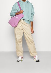 Sudadera con capucha azul claro, pantalones de pierna ancha beige y una bolsa de mano morada con texto blanco; combinada con zapatillas multicolores.