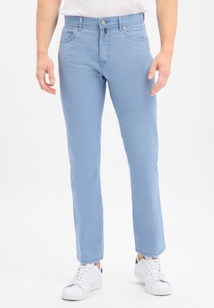 Mann trägt hellblaue Slim-Fit-Jeans und weiße Sneaker, steht vor weißem Hintergrund.