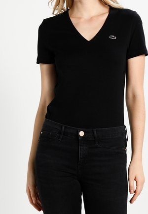 Kobieta ubrana w dopasowaną czarną koszulkę z dekoltem w kształcie litery V i małym białym logo krokodyla na piersi, w połączeniu z czarnymi jeansami o wysokim stanie.