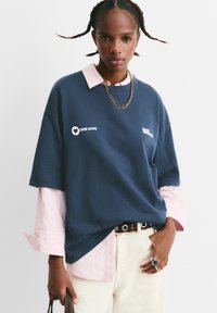 Mørkeblå oversized t-shirt med hvid tekst, lagdelt over en pink stribet skjorte. Har en rund halsudskæring og korte ærmer.