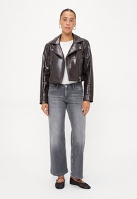 Brun läderkort motorcykeljacka över en vit skjorta, kombinerad med grå vida jeans och svarta ballerinas; modellen står framför en enfärgad bakgrund.