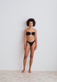 Fekete bikini, vállpánt nélküli felsőrésszel, amelyet díszes ezüst akcentus díszít a középen. Az alsó rész alacsony derekú kialakítású, sima anyagot mutat be.