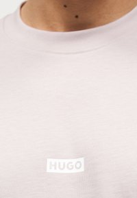 Κοντινή λήψη ενός ανοιχτόχρωμου πουλόβερ με στρογγυλή λαιμόκοψη, με τη λέξη «HUGO» τυπωμένη με ανοιχτό χρώμα στο στήθος.