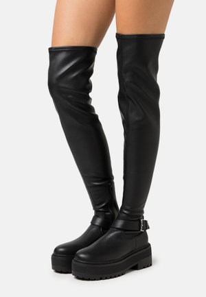 Bottes en cuir noir au-dessus du genou avec une plateforme épaisse et un accent de boucle. Présente une texture lisse et une fermeture éclair sur le côté.
