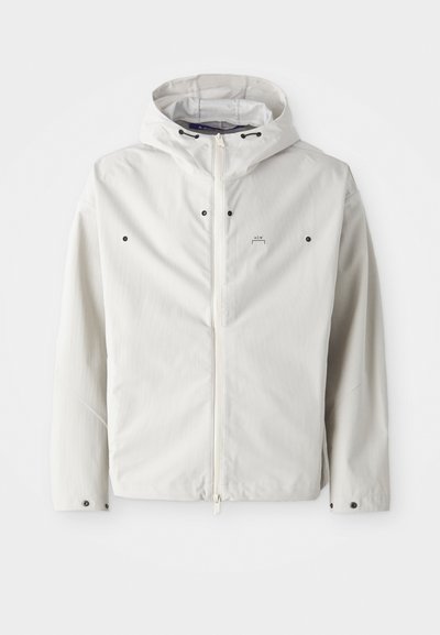 Chaqueta blanca ligera con capucha, cierre de cremallera y botones de acento negros. Textura suave con un sutil patrón de rayas. Branding mínimo.