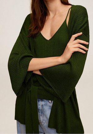 Femme portant un pull en maille côtelée vert foncé à manches larges par-dessus un caraco assorti et un jean bleu clair, bras croisés.