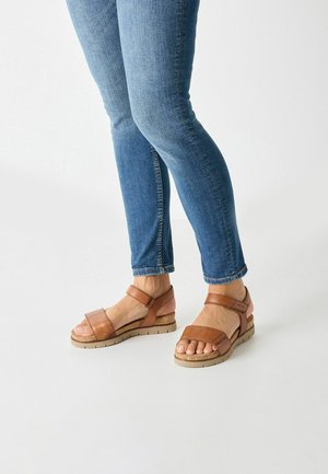 Brown leather sandals with two adjustable straps, a cork wedge sole, and a textured outsole, paired with blue skinny jeans.