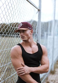 Top nero, cappellino bordeaux con logo, corporatura muscolosa, appoggiato a una recinzione in catena, sfondo neutro con erba.