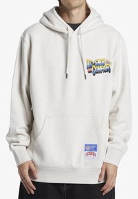 Kremowa bluza z kapturem posiada przednią kieszeń, regulowany sznurek w kapturze oraz haftowane logo na piersi z metką na dole.