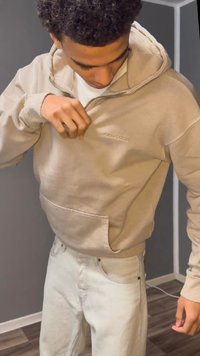 Beige hoodie with a kangaroo pocket, ribbed cuffs, a drawstring hood, and subtle embossed logo. Worn with light-colored jeans.