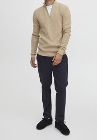 Beżowy pulower z zamkiem do ćwiartki, wykonany z fakturowanego dzianego materiału, w zestawieniu z ciemnoniebieskimi spodniami i czarnymi butami. Stylizacja jest casualowa i dopasowana.