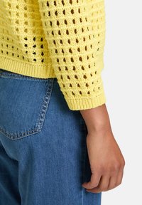 Pull en laine jaune avec un motif ouvert et lâche, muni de poignets côtelés, associé à un jean en denim bleu taille haute.