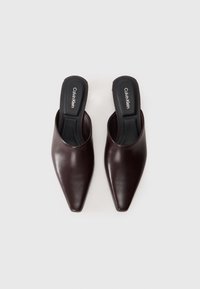 Mule in pelle marrone con design a punta. Presenta una texture liscia e una soletta nera con il logo "Calvin Klein".