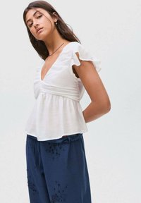 Weißes Kurzarmbluse mit V-Ausschnitt und geraffter Taille. Getragen mit marineblauen Hosen, die bestickte Blumenmuster aufweisen.