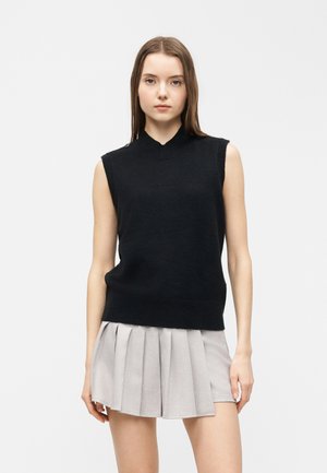 Femme portant un haut noir en maille sans manches avec des détails de boutons et une mini-jupe plissée gris clair, se tenant devant un fond uni.