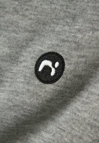 Grå stof med en subtil tekstur har et broderet sort cirkulært logo, der forestiller et stiliseret lille bogstav inden for cirklen.