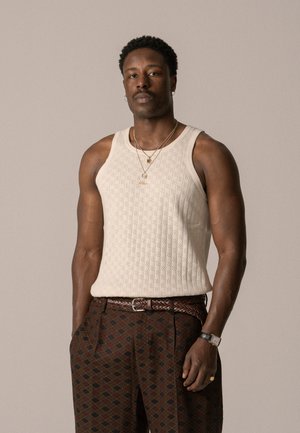 Beige knitted tank top with textured pattern, paired with brown patterned pants featuring a diamond design and a braided brown belt.