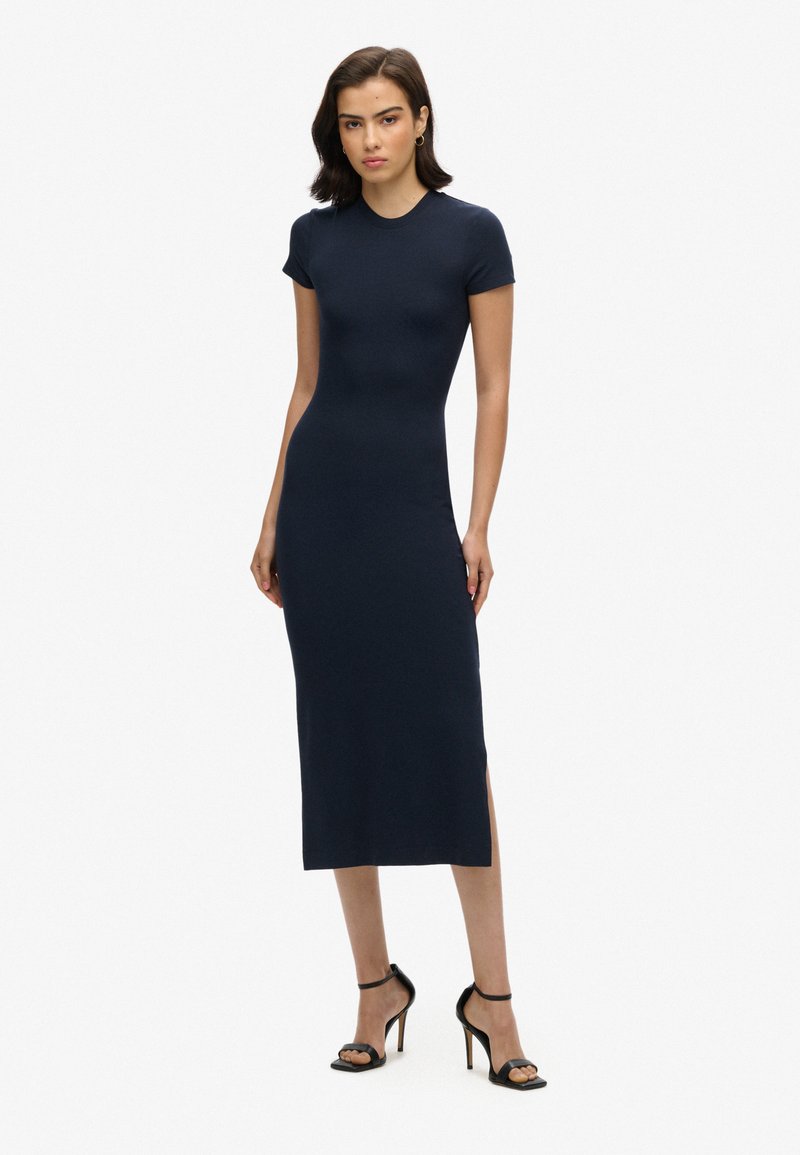 Robe midi ajustée marine avec un col rond, des manches courtes, une fente sur le côté et une texture lisse. Associée à des sandales à talons noires.