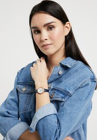 Montre pour femme avec un cadran blanc et des accents dorés sur un bracelet en cuir bleu marine, porté sur un poignet, sur fond de veste en denim.