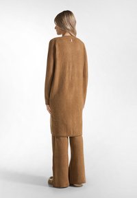 Lange bruine gebreide trui met een ronde halslijn, zijspleten en een relaxte pasvorm, gecombineerd met bijpassende wijde broek. Gelaagd, zacht materiaal.