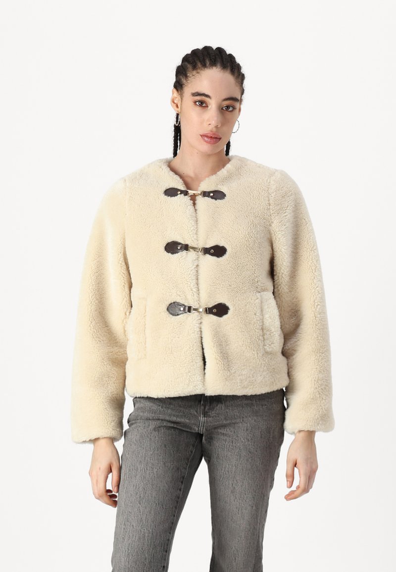 Giacca in pelliccia sintetica color crema con scollatura rotonda, dotata di chiusure a toggle marroni e due tasche frontali. Texture morbida, vestibilità rilassata.