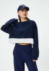Sudadera navy recortada con un dobladillo blanco en capas y detalles de botones. Combinada con pantalones navy de cintura elástica, completada con una gorra a juego.