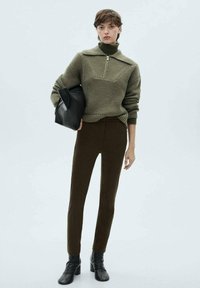 Maglione lavorato a maglia in verde oliva con colletto alto e zip, abbinato a pantaloni bruniti aderenti e stivali neri alla caviglia. Il modello tiene in mano una borsa nera.