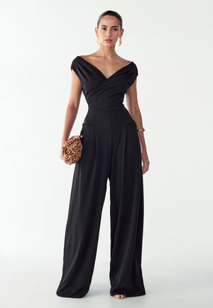 Sort jumpsuit med indpakket off-shoulder halsudskæring, vide ben og sidelommer. Kombineret med en tekstureret clutch med gulddetaljer.