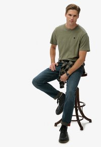 Krótki rękaw t-shirt w oliwkowej zieleni z logo, niebieskie dżinsy, koszula w kratę przewiązana w talii, czarne sznurowane buty oraz zegarek na nadgarstku, siedząca na stołku.