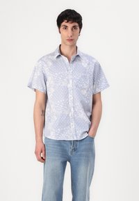 Camicia a maniche corte, di colore azzurro chiaro, con motivi floreali bianchi e a pois, colletto con bottoni e taschino frontale. Jeans in denim con gamba dritta.