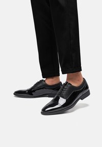 Scarpe eleganti nere con finitura in pelle verniciata e dettagli in rete sui lati, progettate con punta arrotondata e tacco basso.