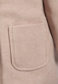 Tkanina za svetlo roza volneno plašč, ki vključuje zaobljen, našit žep z mehko teksturo in čistimi linijami, kar poudarja minimalistični dizajn.