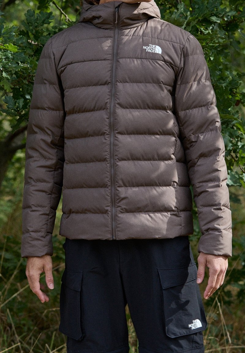 Bruine gewatteerde jas met ritssluiting en capuchon, voorzien van een doorgestikt ontwerp. Opvallend logo op de linkerborst. Gecombineerd met zwarte cargobroeken.